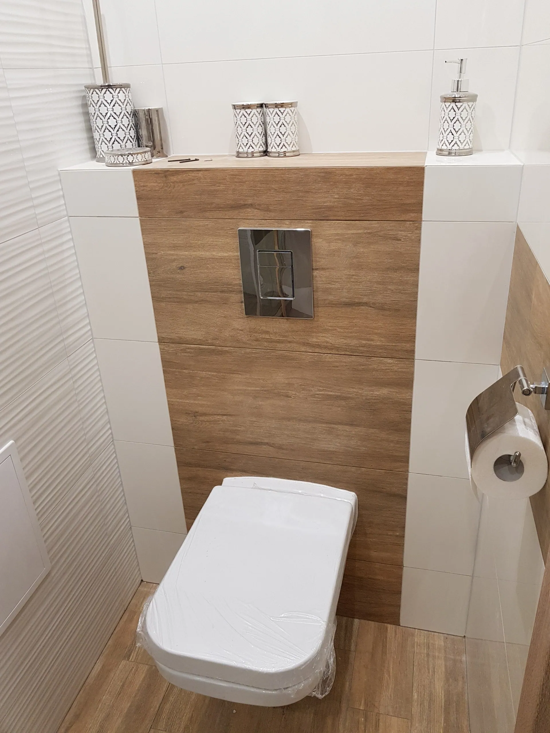 Wykończone WC z zabudową stelaża podtynkowego obłożoną gresem drewnopodobnym i biała płytka strukturalna na ścianach.